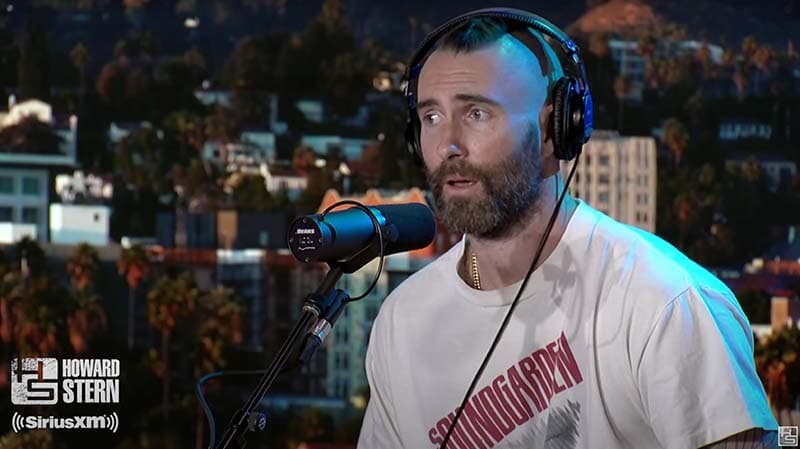 Image shows the Adam Levine(a white man, with dark hair and a dark beard), wearing a white t-shirt sitting and sitting in a studio. He is speaking in to a SHURE SM7B microphone.