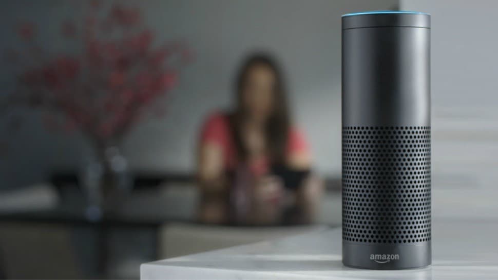 Image shows a black, cylindrical, Amazon smart speaker on a table top. In the blurred background a woman is sitting at a table and using her phone.