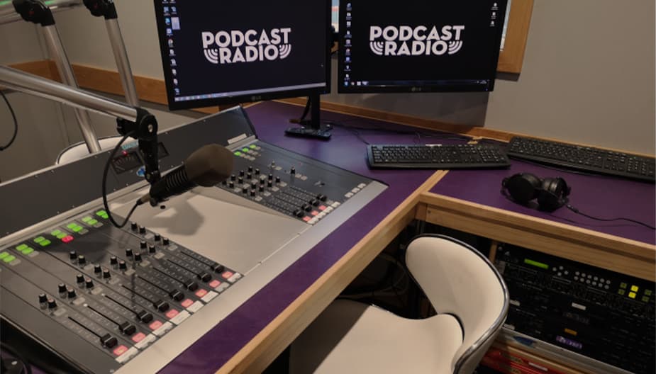 A radio studio with 2 computer screens, a mixing desk and a microphone.