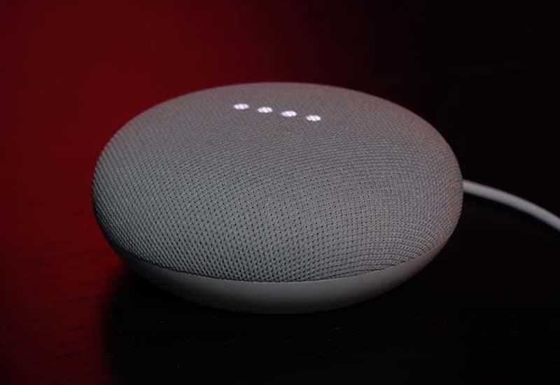 Image shows a grey Google Nest Mini smart speaker, on a table with a power cable plugged into it.