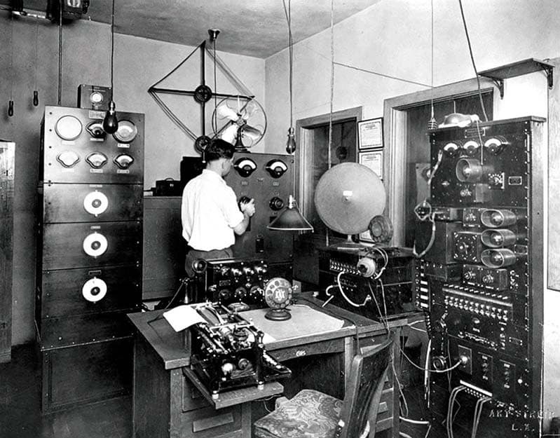 A black and white image of the KNX Transmitter, circa 1926. A man with his back to the camera is adjusting a setting on the transmitter.