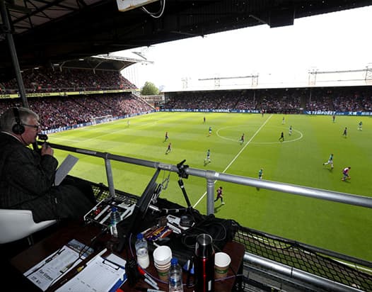 A commentator, reporting on a live game from a broadcasting desk in a football stadium. The commentator and desk are overlooking the pitch.