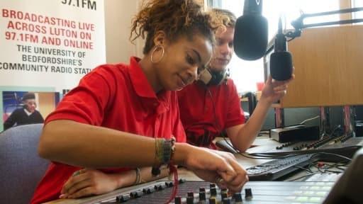 A man and a woman sitting side by side in a radio booth. The woman is putting down the fader on the mixing desk.