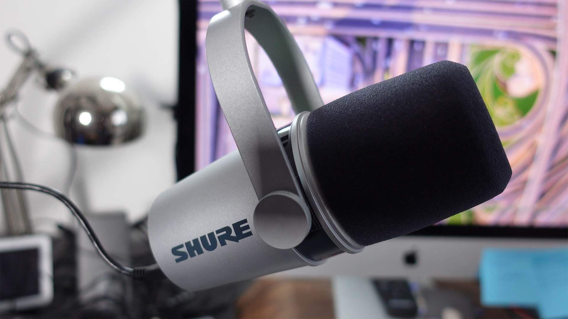 Image shows a silver Shure MV7, side on and hanging from a boom arm. In the background is a computer and desk lamp.