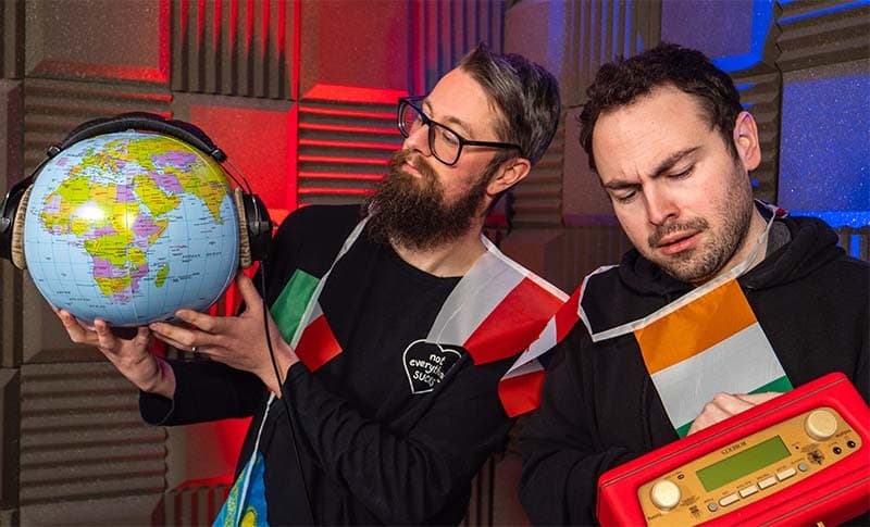 Two men with flag bunting around their shoulders, one is holding an inflatable globe with headphones on it, the other is holding a red Roberts radio.