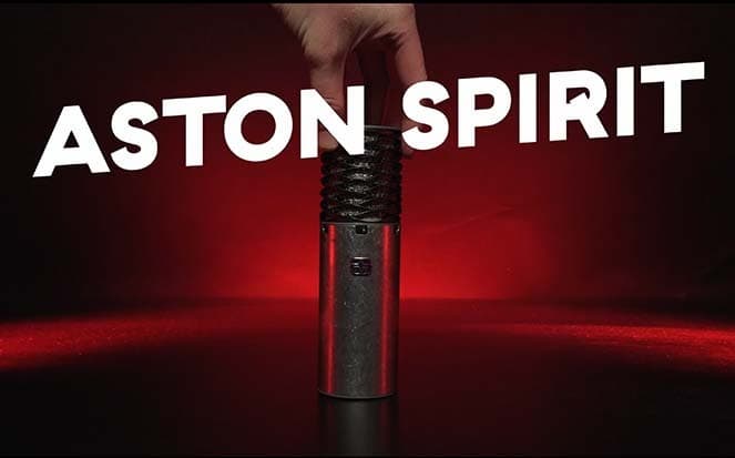 Image shows an Aston Spirit microphone, being stood up on a table by someone holding the top end. The microphone is silver metal and cylindrical shaped. It's lit up by a red spotlight, against a dark background. The image also has white, capitalised text running across the image that reads: ASTON SPIRIT.