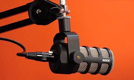 Image shows the black Rode PodMic, suspended by a mic stand and shock mount. The background is a block orange colour.