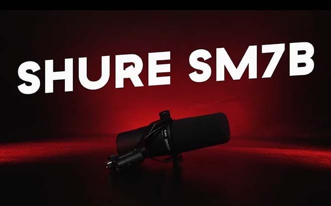 Image shows the black Shure SM7B placed on a table, lit with a red spotlight against a dark background. There is white, capitalised text that is slanted diagonally across the image which reads: SHURE SM7B