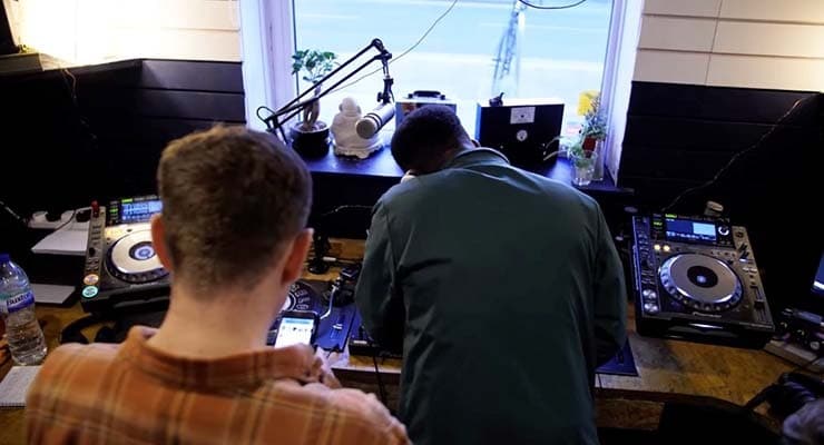 Two men in a radio booth. One man is mixing on the decks and the other is standing behind him and playing on his phone.