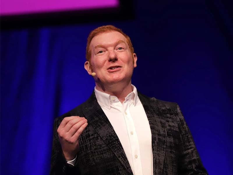 Image shows a white man with red hair and wearing a black suit jacket and white shirt, delivering a speech on a stage.